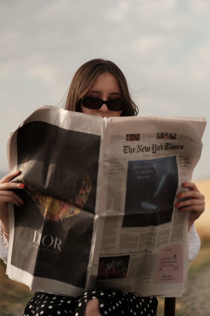 An adult woman enjoys reading The New York Times while sitting outdoors on a sunny day.