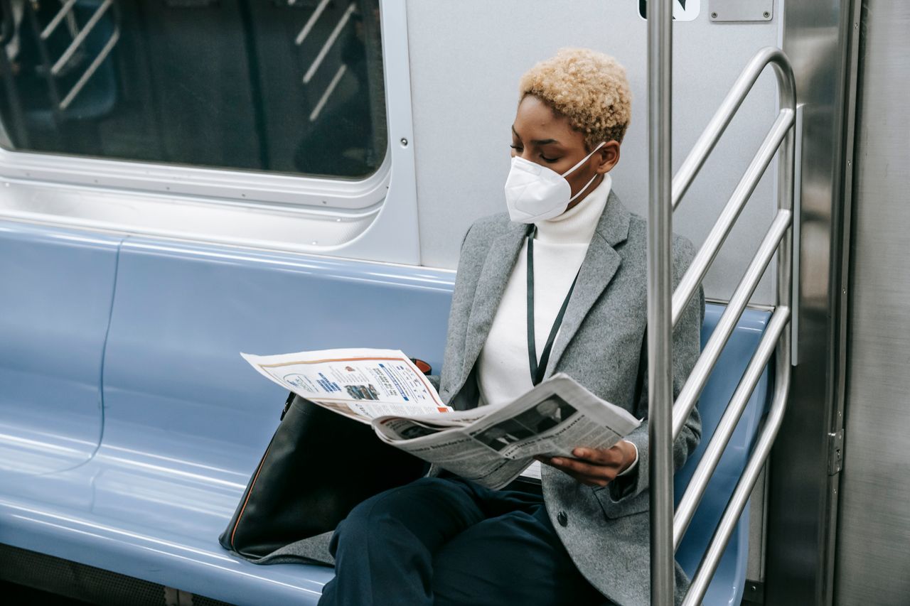 Unrecognizable concentrated young African American female manager with short dyed hair in elegant outfit and face mask commuting on subway train and reading newspaper during COVID 19