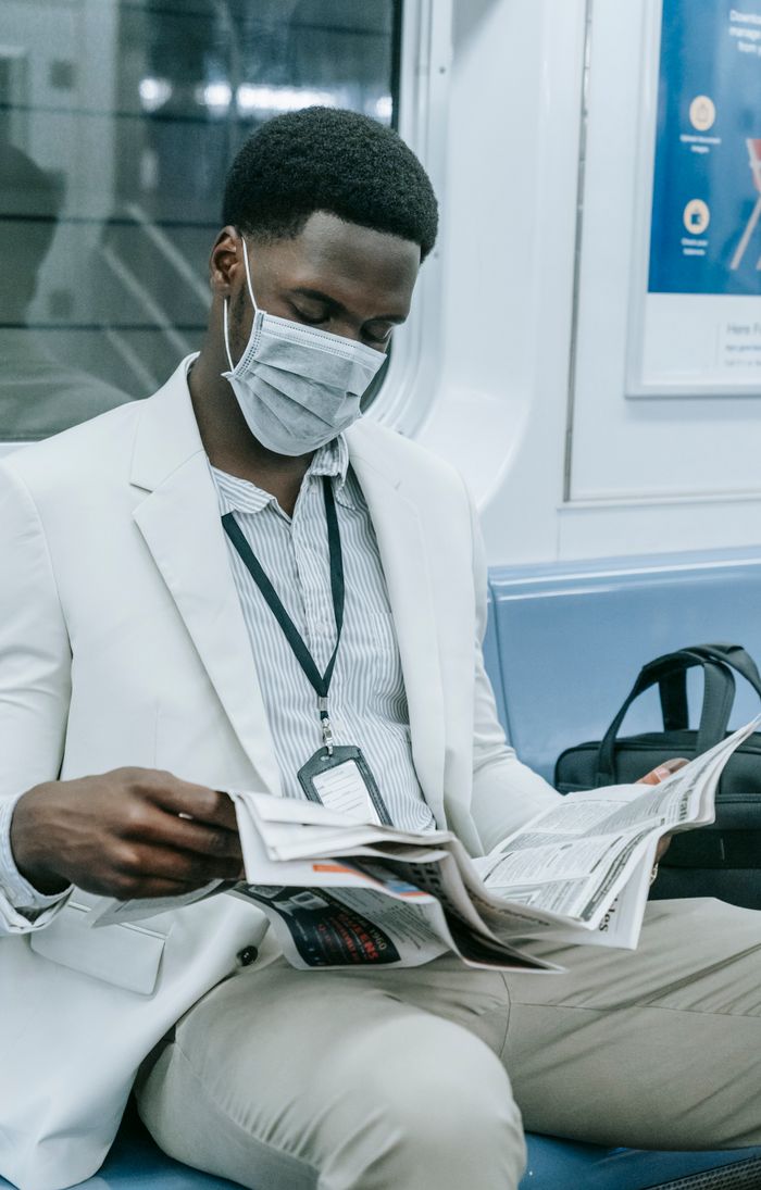 A man wearing a mask and suit reads a newspaper on a train, capturing urban commuting during the pandemic.